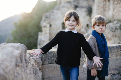 Portrait of happy siblings standing by retaining wall