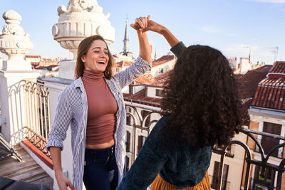 Excited young multiracial female friends wearing casual outfits holding hands and dancing together while spending sunny day on building rooftop