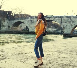 Full length portrait of young woman standing against bridge