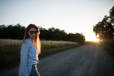 Portrait of young woman standing by road against clear sky