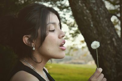 Side view of young woman blowing dandelion seed while standing in park
