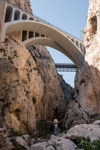 Back view of distant anonymous female traveler standing with spread arms while admiring aged bridges in mountainous area during trip in spain