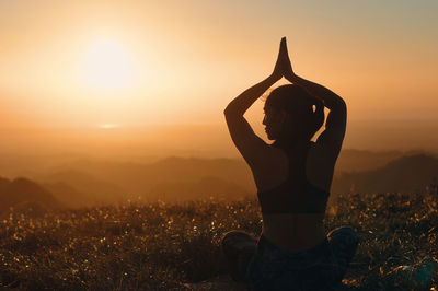 Rear view of woman meditating on field against sky during sunset