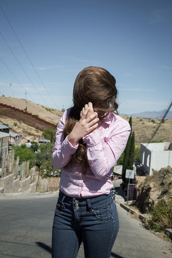 Woman covering face with hair on street against sky during sunny day