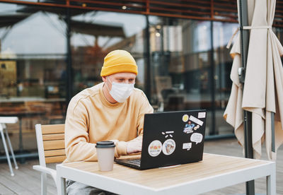 Stylish man hipster in face mask freelancer working on laptop in street cafe, new normal