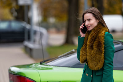 A woman talking on the phone while standing by a green car parked on a city street in the background