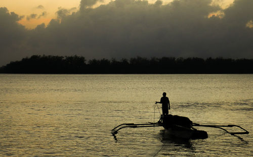 Silhouette fisherman sailing outrigger in lake against cloudy sky