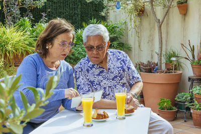 Senior woman showing digital tablet to man while having breakfast while sitting in yard