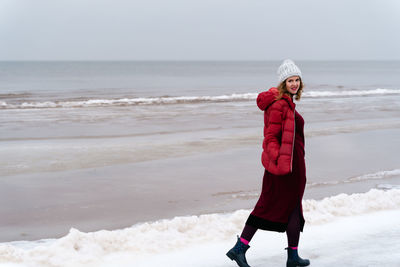 A woman in a red jacket and a red knitted dress walks in winter along the sea