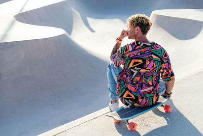 High angle back view of trendy tattooed millennial guy with curly hair wearing stylish colorful shirt and jeans sitting on skateboard near ramp in skatepark
