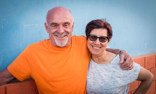 Portrait of smiling senior couple with arm around standing against wall