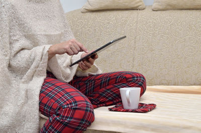 Woman in cozy home wear sitting on sofa, having coffee, holding tablet and reading