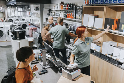 Female sales clerk showing various appliances on shelf to customer at checkout counter