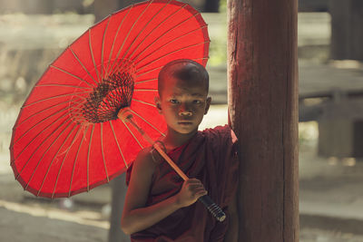 Portrait of monk holding red umbrella by wooden column