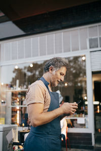 Side view of male owner using mobile phone outside deli