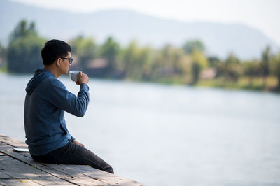 Side view of man drinking coffee while sitting on pier over lake