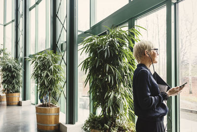 Mature businesswoman looking through window while holding smart phone in office