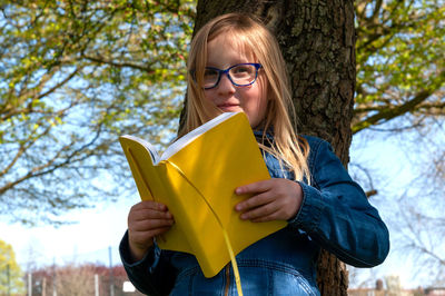 Young school age girl reading from yellow textbook in park.