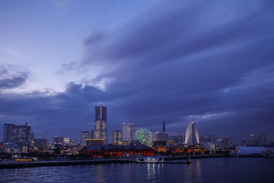 Illuminated city by river against sky at dusk