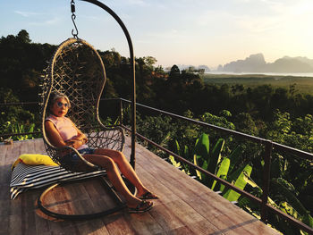 Woman sitting on hammock by railing against sky