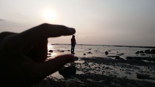 Optical illusion of silhouette hand holding woman standing at beach against sky during sunset