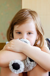 Dramatic portrait of little girl sitting on chair and cuddling teddy bear with sad face.