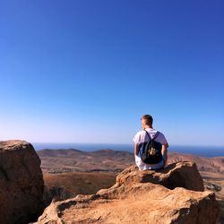 Rear view of man sitting on rock against sky