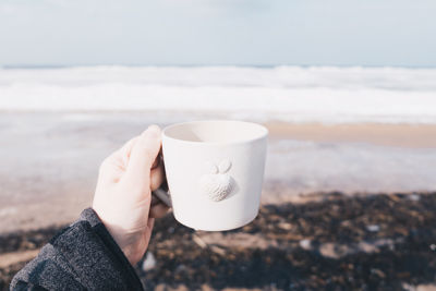 Cropped hand holding coffee cup at beach against sky