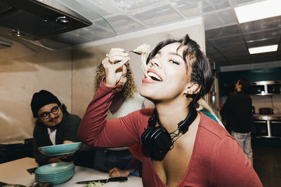 Happy young woman eating noodles by friend in kitchen at college dorm