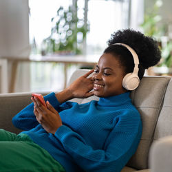 Happy african american woman in headphones listening to music using mobile phone lies on sofa