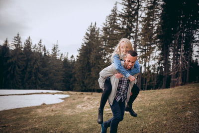 Happy man piggybacking girlfriend on field against trees