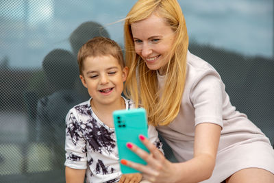 Happy blond woman and little boy sitting on terrace and making selfie on smartphone. mother and son