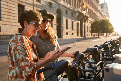 Happy young girlfriends using smartphone to rent electric scooter on sunlit street of madrid at sunset in spain