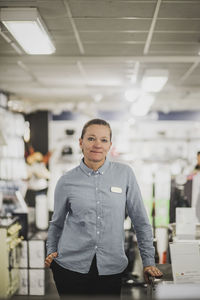 Portrait of smiling mature saleswoman with hand in pocket standing in electronics store