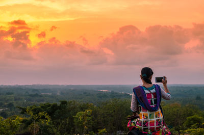 Rear view of woman photographing on mobile phone against sky during sunset