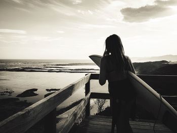 Rear view of woman with surfboard standing at beach against sky