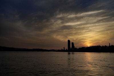 Silhouette of buildings by sea against cloudy sky