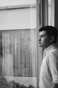 Side view of thoughtful young man looking through window at home