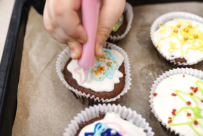 A child squeezes colored frosting from a tube onto chocolate brown cupcakes covered white frosting.