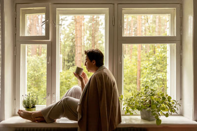 Middle age beautiful woman sitting on windowsill, drinking tea, dreaming. 50-year-old woman relaxing