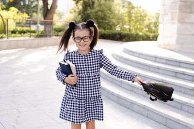 Pretty little child girl 5-6 year old playing outdoors wear glasses, stylish dress holding backpack.