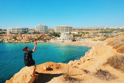 Hiker sitting on rock at lakeshore by city against clear blue sky