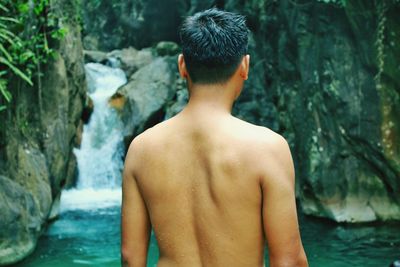 Rear view of shirtless man standing against waterfall