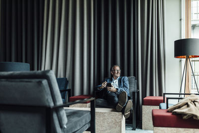 Smiling businessman holding mobile phone while relaxing in cafeteria at office