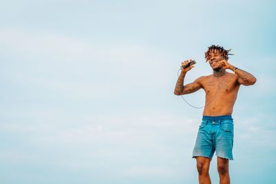 Full length of shirtless man listening to music against sky