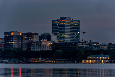 Illuminated buildings by river against sky at dusk