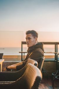 Side view of young man looking away while sitting on chair against clear sky during sunset