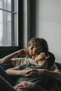 Thoughtful lesbian woman looking through window while lying down with girlfriend on sofa at home