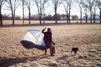 Full length of man carrying portable tent while walking with dog on field