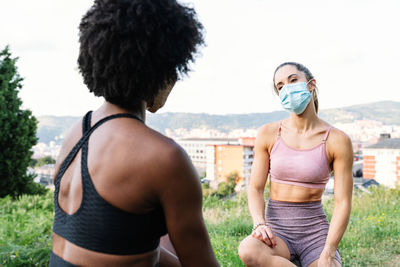 Young fit female friends in activewear and protective masks sitting on wooden fence and chatting while having break during outdoor training in summer day